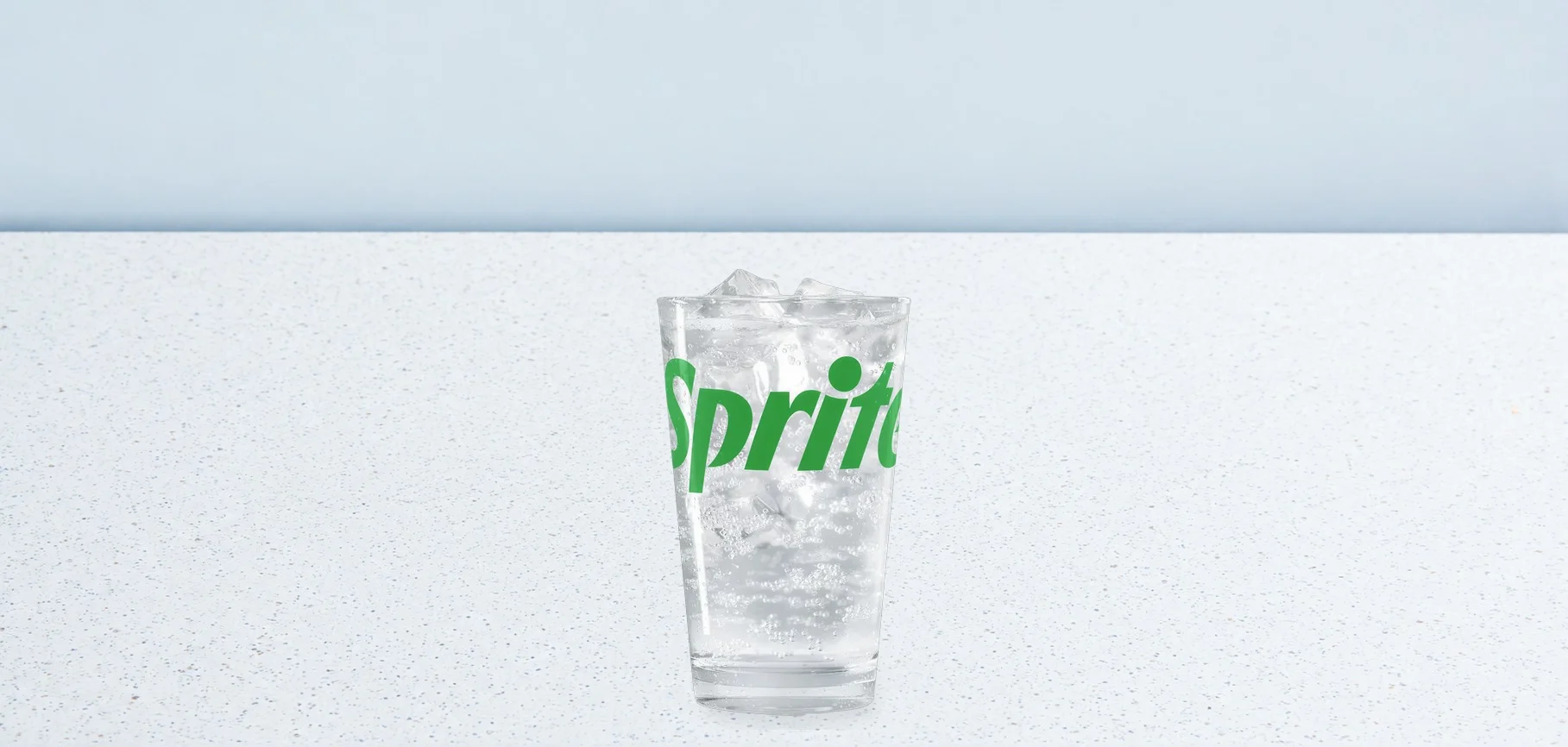 Glass of Sprite with ice cubes on a white speckled countertop, light blue background, green Sprite logo visible on the glass.