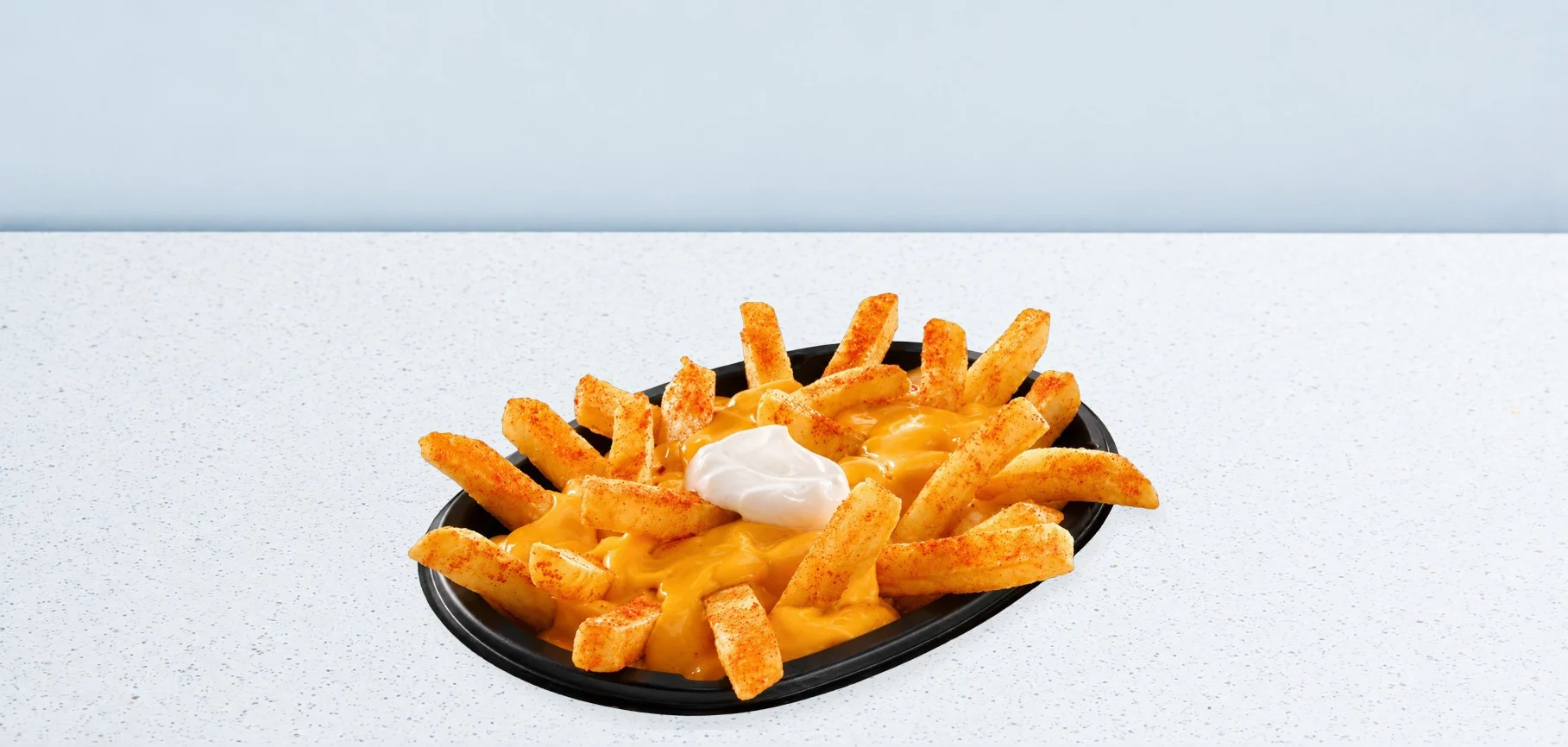 Plate of cheese fries sprinkled with paprika, with a dollop of sour cream on a white speckled table.