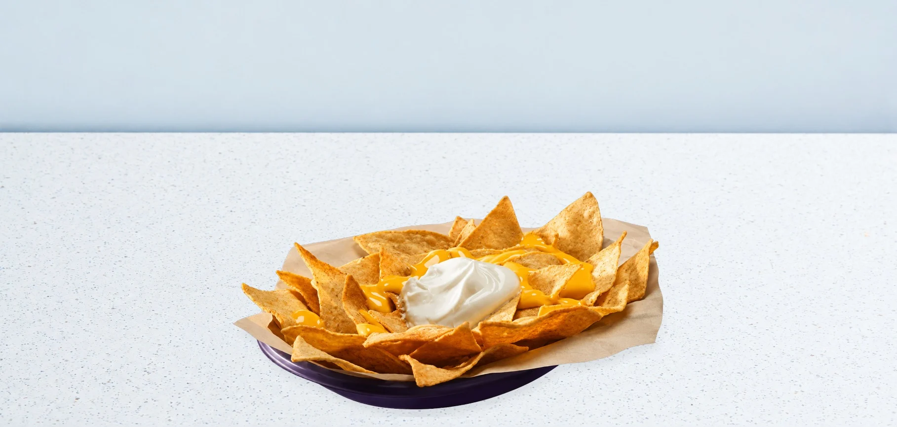 3411-cheesy-nachos | Taco Bell Hrvatska Tortilla chips on a purple plate with melted cheese and a scoop of sour cream on parchment, set on a pale speckled table.