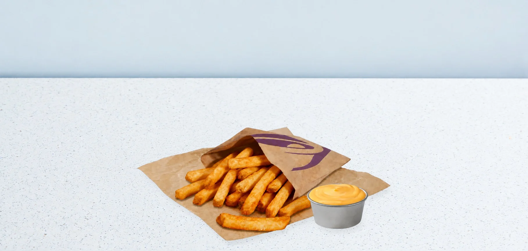 3413-xl-fries-nacho | Taco Bell Hrvatska French fries spilling from a brown paper bag with a purple logo, beside a cup of yellow cheese sauce on a pale speckled counter surface.